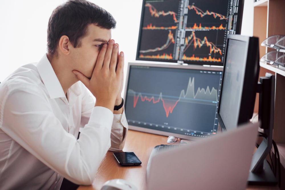 Stressed male trader in a white shirt sitting at a desk, holding his face in his hands while analyzing financial charts on multiple computer monitors showing market downturns.