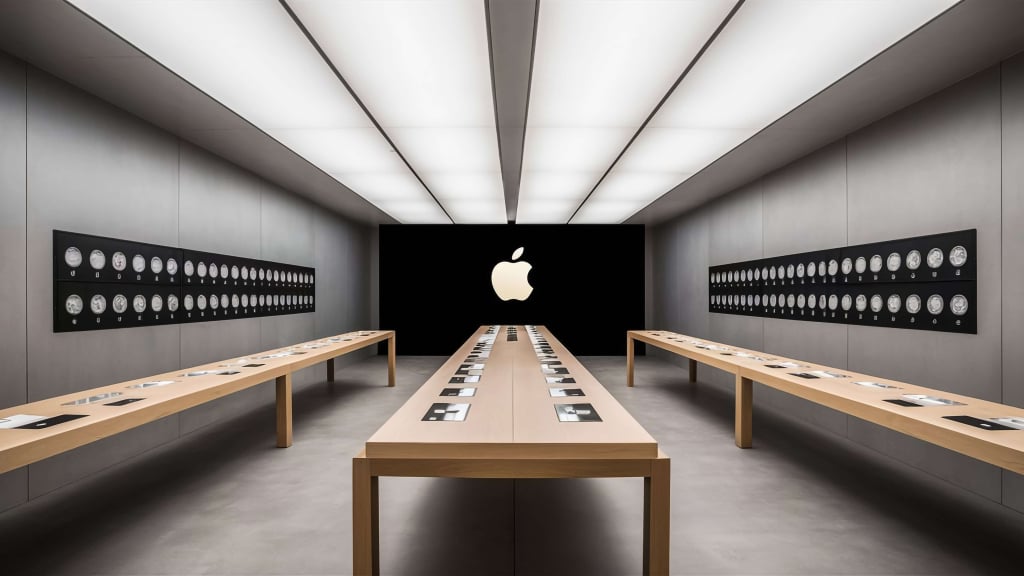 Modern Apple Store interior with minimalist design, showcasing light wood display tables, wall-mounted Apple Watch displays, and a large illuminated Apple logo on a black wall.