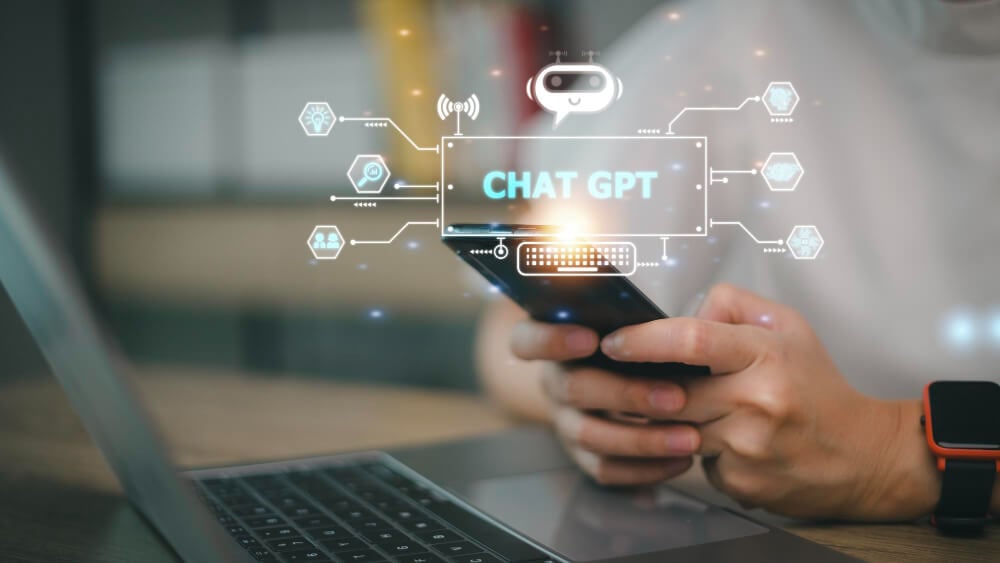 Person holding a smartphone with a digital overlay that reads ‘Chat GPT,’ surrounded by icons representing AI and technology, sitting next to an open laptop.