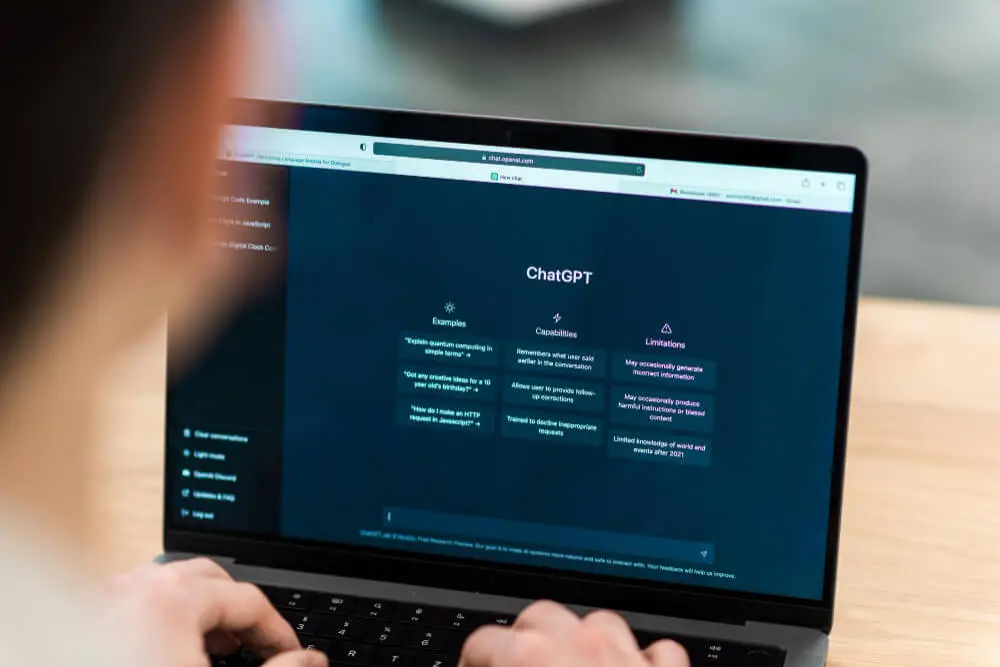 A person using a laptop with the ChatGPT interface open on the screen, displaying sections for examples, capabilities, and limitations.